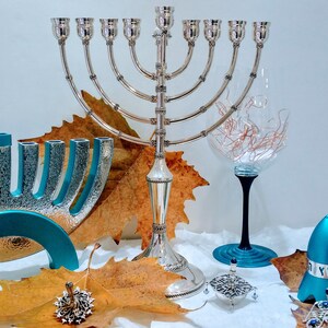 May include: A silver menorah with nine candlesticks, a blue menorah with nine candlesticks, two blue dreidels, a silver dreidel, and a wine glass with a black stem and a blue base. The menorahs and dreidels are surrounded by autumn leaves.