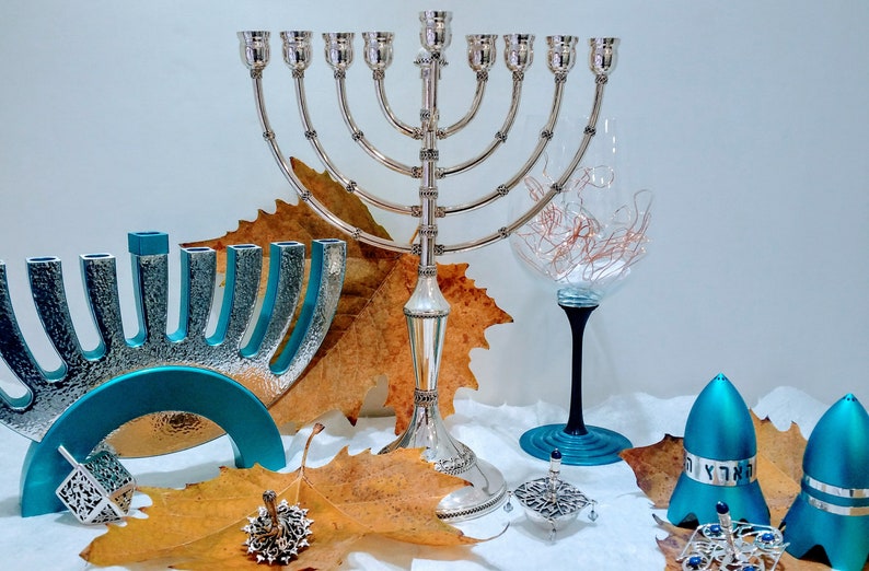 May include: A silver menorah with nine candlesticks, a blue menorah with nine candlesticks, two blue dreidels, a silver dreidel, and a wine glass with a black stem and a blue base. The menorahs and dreidels are surrounded by autumn leaves.
