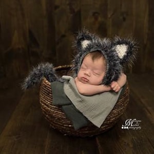 May include: A newborn baby wearing a grey and white crocheted wolf hat with a matching tail, sleeping in a brown wicker basket.