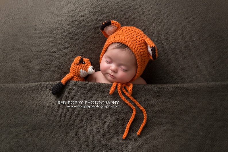 May include: A newborn baby wearing a crocheted orange fox hat with black ears and a matching orange fox stuffed animal. The baby is sleeping on a brown textured fabric.