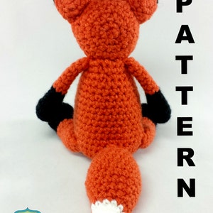 May include: A crocheted fox amigurumi toy with orange fur, black ears and paws, and a white tipped tail. The toy is sitting on a white surface with the word "PATTERN" written vertically in black letters on the right side of the image.
