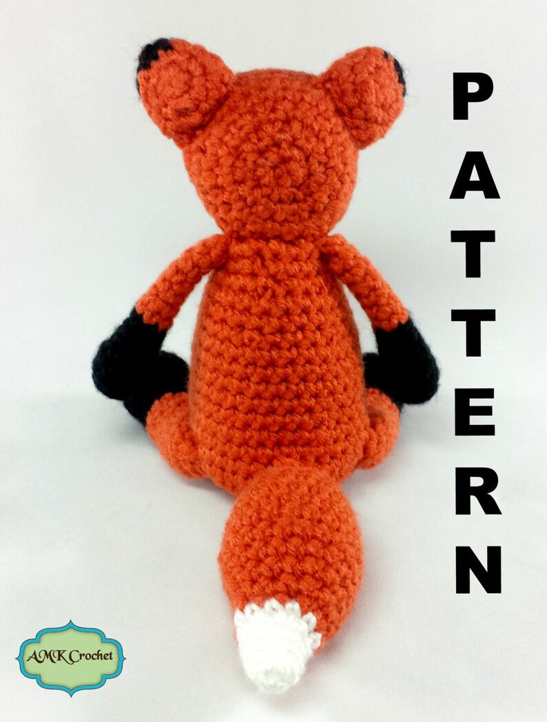 May include: A crocheted fox amigurumi toy with orange fur, black ears and paws, and a white tipped tail. The toy is sitting on a white surface with the word "PATTERN" written vertically in black letters on the right side of the image.