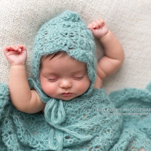 May include: A baby wearing a light blue crocheted hat and matching blanket. The baby is sleeping peacefully with arms outstretched.
