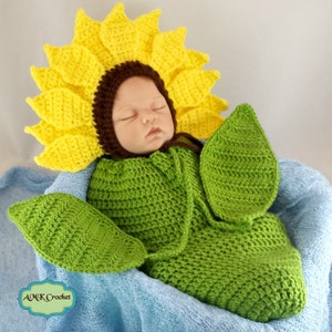 Puede incluir: Un conjunto de bebé de ganchillo en forma de girasol con un gorro a juego. El conjunto es verde y amarillo y presenta un diseño de girasol. El bebé está durmiendo plácidamente en el conjunto.
