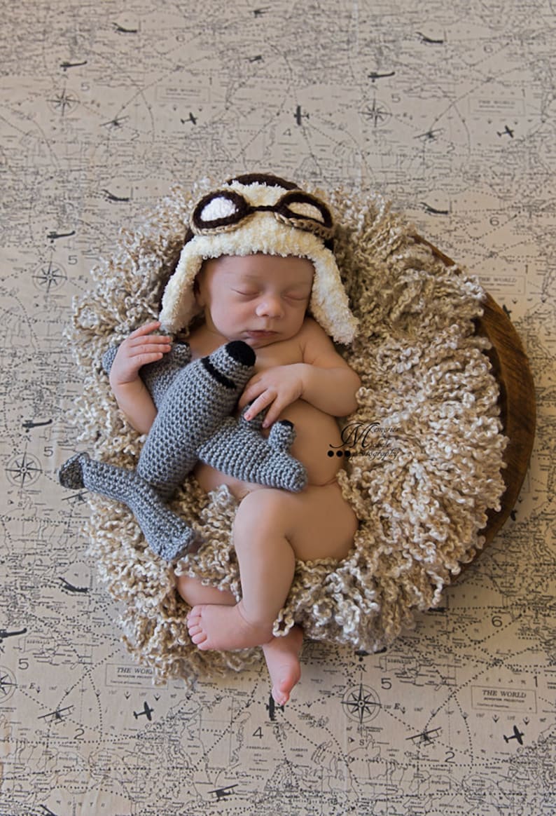 May include: A newborn baby wearing a brown and white crocheted aviator hat with goggles, sleeping in a brown wicker basket on a beige fabric with a world map pattern. The baby is holding a grey crocheted airplane toy.