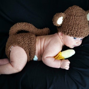 Op de afbeelding: Een baby die een bruin apenpak draagt met een witte staart en een gele banaan als rekwisiet. De baby ligt op een zwart oppervlak.