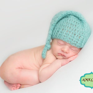 May include: A sleeping baby wearing a light blue crocheted hat with a long pointed tip. The baby is lying on a white blanket.