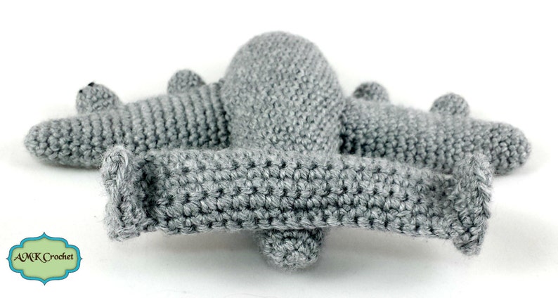 May include: A grey crocheted airplane toy with a long wingspan and a rounded body. The airplane is made with a single crochet stitch and has a textured surface.
