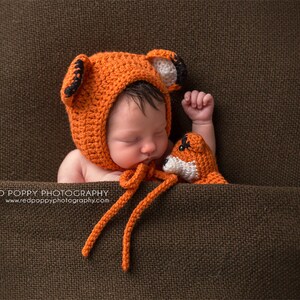 May include: A newborn baby wearing an orange crocheted fox hat and holding a small orange crocheted fox toy. The baby is lying on a brown fabric background.