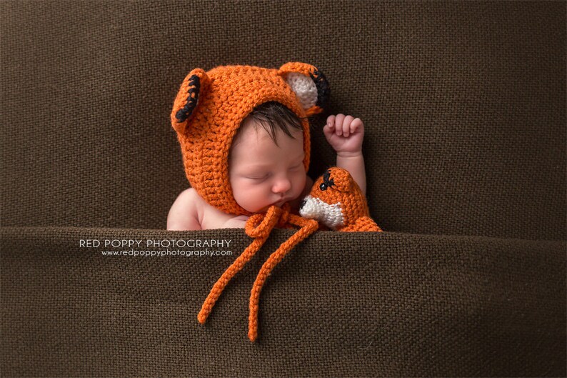 May include: A newborn baby wearing an orange crocheted fox hat and holding a small orange crocheted fox toy. The baby is lying on a brown fabric background.