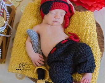 May include: A baby dressed as a pirate is sleeping on a yellow blanket. The baby is wearing a red crocheted hat with a black eye patch, black crocheted pants, and is holding a gray crocheted sword. There are gold coins scattered around the baby.