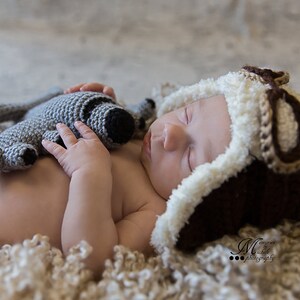 May include: A newborn baby wearing a brown and white crocheted aviator hat sleeps on a white fluffy blanket while holding a grey crocheted stuffed animal.