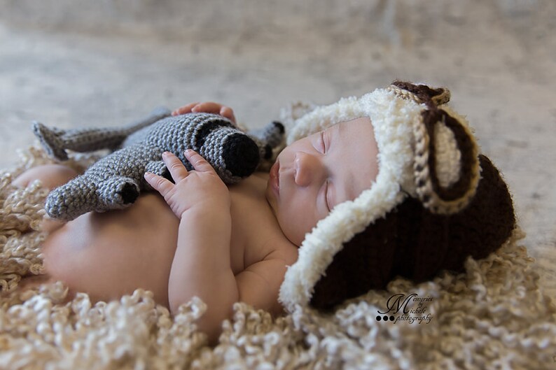 May include: A newborn baby wearing a brown and white crocheted aviator hat sleeps on a white fluffy blanket while holding a grey crocheted stuffed animal.