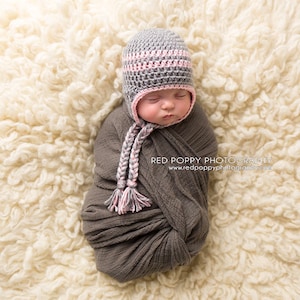 May include: A newborn baby wearing a gray and pink crocheted hat with a long tassel. The baby is swaddled in a gray fabric and is lying on a white fluffy surface.