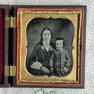 May include: Antique daguerreotype portrait of a woman and child in a hinged case. The case is open, revealing a velvet interior with an embroidered design. The portrait is in a gold-colored frame.
