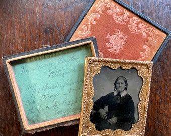 May include: A vintage photograph of a woman in a dark dress, framed in a gold-colored frame. The frame has a decorative design and is sitting on a wooden surface. The photo is labeled with handwritten text: "Mo Nemy Loy the Pittofurce 14/17 Bust-Remove Cap Stringo-Brut The fur Qual Way"