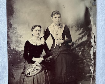 Tintype of two well dressed women (sisters?) posed before a painted backdrop.