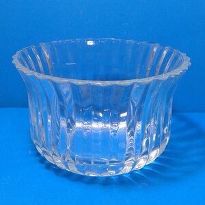 Puede incluir: Un cuenco de vidrio transparente con un diseño acanalado. El cuenco tiene un borde ancho y festoneado y una base redondeada. El patrón acanalado crea líneas verticales. El cuenco está sobre una superficie azul.