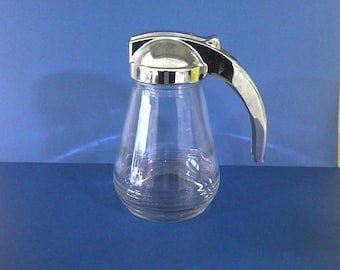 May include: A clear glass syrup dispenser with a chrome-colored lid and handle. The dispenser has a rounded base and a tapered body. The handle curves upward, and the lid has a hinged design. The background is a solid blue.
