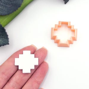 May include: Two pixelated cross-shaped objects. One is white and held in a hand, the other is peach-coloured and sits on a white surface. The objects are likely cookie cutters or craft supplies.