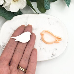 Könnte beinhalten: Ein weißer Plastik-Ausstecher in Form eines Vogels und ein rosa Plastik-Ausstecher in Form eines Vogels auf einer weißen Marmorfläche.