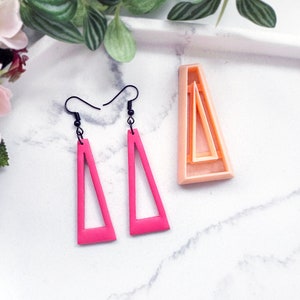 Peut inclure: Une paire de boucles d'oreilles triangulaires roses avec des crochets noirs. Les boucles d'oreilles sont en argile polymère et ont un centre creux. Les boucles d'oreilles sont présentées à côté d'un moule triangulaire rose.