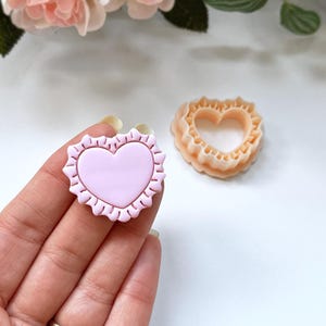 Könnte beinhalten: Ein hellrosa herzförmiger Ausstecher mit gewelltem Rand wird in einer Hand gehalten. Daneben befindet sich ein pfirsichfarbener herzförmiger Ausstecher. Die Ausstecher befinden sich auf einer weißen Oberfläche mit Blumen im Hintergrund.
