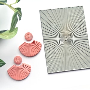 Puede incluir: Un par de pendientes en forma de abanico de color coral con una superficie texturizada, junto a un sello rectangular gris con un diseño de rayos de sol. Los pendientes tienen una parte superior circular y una parte inferior curvada y acanalada.