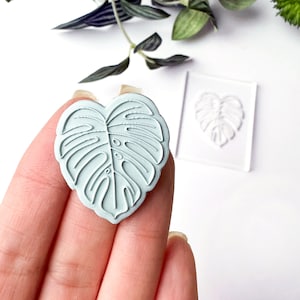 Puede incluir: Un molde de silicona azul claro con forma de hoja de monstera. El molde está en una mano, y un molde de silicona transparente de la misma hoja está en el fondo.