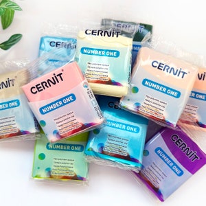 May include: A close-up of a pile of colourful polymer clay bars, each labelled "Cernit Number One". The bars are packaged in clear plastic wrappers and feature instructions for baking the clay at 110-130 degrees Celsius for 10 minutes or 230-265 degrees Fahrenheit for 30 minutes.