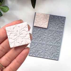 May include: A silicone mold with six square impressions of a floral design. The mold is gray and the impressions are raised. A pink silicone square with the same floral design is shown next to the mold.