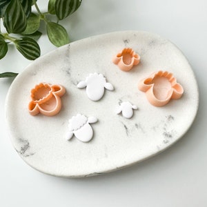 May include: A collection of sheep-shaped cookie cutters and fondant sheep decorations are displayed on a white, oval platter. The cutters are peach-coloured, and the fondant sheep are white. The platter has a marble-effect pattern.