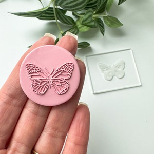 May include: A pink silicone stamp featuring a detailed butterfly design. The stamp is held in a hand, with a clear acrylic stamp showing the same butterfly design in the background.