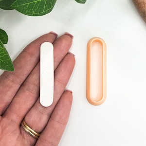 Può includere: Uno stampo rettangolare bianco con un bordo arrotondato e uno stampo in silicone abbinato di colore pesca. Entrambi gli stampi sono progettati per creare una forma rettangolare lunga e sottile.