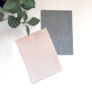 May include: Two textured rectangular placemats, one in a light pink color and the other in a dark gray color.