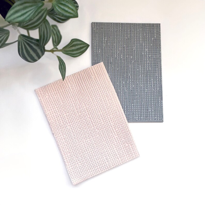 May include: Two textured rectangular placemats, one in a light pink color and the other in a dark gray color.