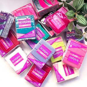 May include: A collection of rectangular Sculpey Soufflé oven-bake clay blocks in various colours including pink, teal, purple, lime green, white, and red. Each individually wrapped package features the Sculpey Soufflé logo and weighs 48 g.