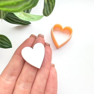 May include: Two heart-shaped cookie cutters, one white and one peach. The white cookie cutter is being held in a hand.