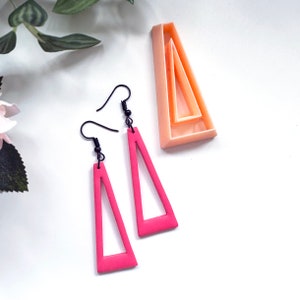 Peut inclure: Une paire de boucles d'oreilles triangulaires roses avec un centre creux. Les boucles d'oreilles sont en argile polymère et ont des crochets en métal noir.
