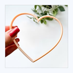 May include: A light peach-coloured heart-shaped cookie cutter with a thin, open frame design. The object is constructed from a smooth, solid material and features a classic, symmetrical heart outline with a pointed bottom and two rounded top lobes.