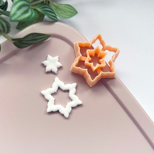 May include: Three white fondant snowflake shapes on a light pink surface. A larger orange plastic snowflake cookie cutter is also on the surface.