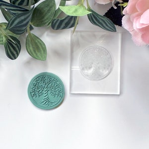 Peut inclure: Un tampon en acrylique transparent avec un motif d'arbre de vie. Le tampon est sur une surface blanche à côté d'un tampon en caoutchouc vert sarcelle avec le même motif.