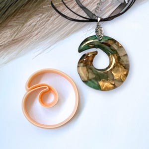 Pode incluir: Um colar com um pingente de espiral verde e dourado com um fecho prateado. O pingente é feito de resina e tem um design único e abstrato. O colar está em um cordão preto.