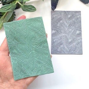 May include: Two rectangular stamps with a palm leaf pattern. One is light green, held in a hand, and the other is grey. The stamps feature a raised, textured design of overlapping palm leaves.