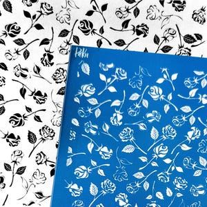 May include: A blue stencil featuring white rose designs and the word "Paku". A white fabric with black rose patterns is in the background. The roses have detailed petals and stems.