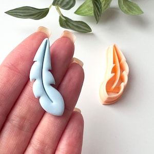 Könnte beinhalten: Zwei blaue und orangefarbene Plastik-Ausstecher in Form von Federn. Der blaue Feder-Ausstecher wird in einer Hand gehalten.