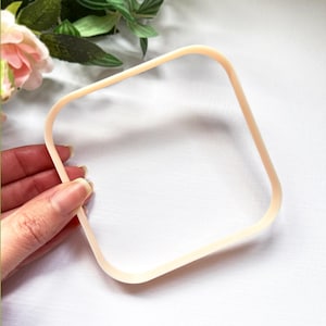 May include: A light beige, square-shaped frame with rounded corners. The frame is constructed from a smooth, solid material and features a simple, open design. It is held in a position that displays its clear, hollow center and uniform thickness.