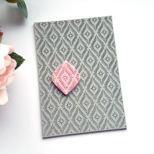 May include: A gray silicone stamping block with a repeating geometric pattern. A smaller pink silicone stamp with a similar pattern is placed on top of the block.