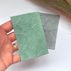 May include: Two rectangular, textured leaf-patterned tiles. One is a soft green, the other a muted gray. The leaf design is raised, creating a tactile surface. The tiles are likely for decorative or craft purposes.
