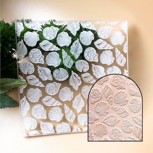 Op de afbeelding: Een transparante acryl stempel met een reliëf ontwerp van schelpen. De stempel wordt getoond op een lichtbruine ondergrond met een close-up van het ontwerp dat is geëmbost op een roze oppervlak.